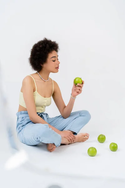 Молодая африканская американка в верхней части держит зеленое яблоко, сидя на сером фоне — стоковое фото