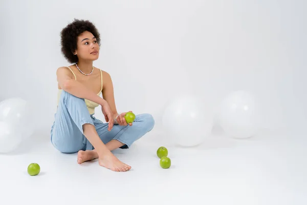 Mujer afroamericana descalza sosteniendo manzana cerca de globos sobre fondo gris - foto de stock