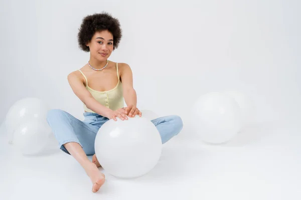 Barefoot african american woman looking at camera near balloons on grey background — Stock Photo