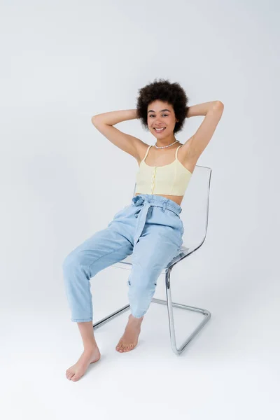 Sonriendo descalza mujer afroamericana sentada en silla sobre fondo gris - foto de stock