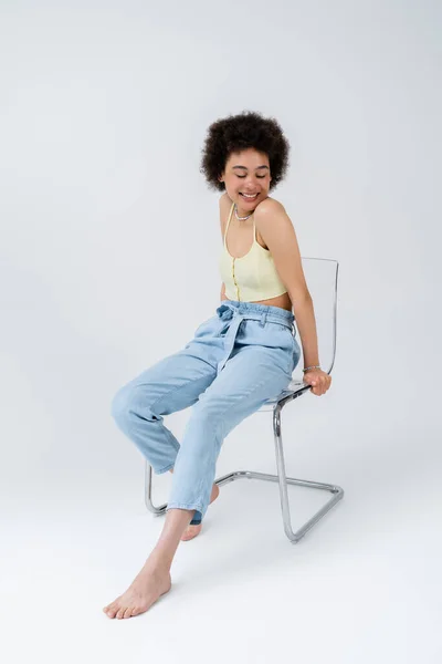 Довольная молодая африканская американка, сидящая на стуле на сером фоне — стоковое фото
