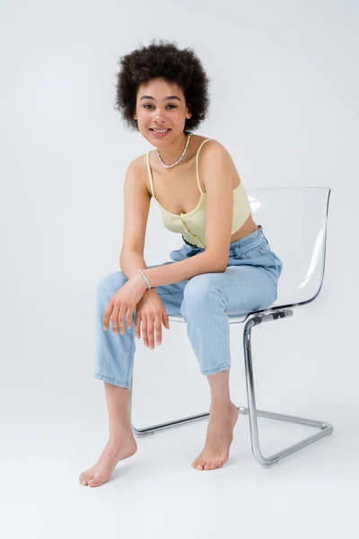 Cheerful young african american woman sitting on modern chair on grey background — Stock Photo