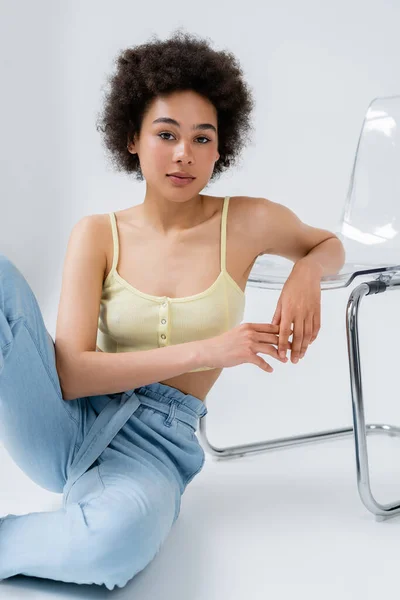 Young african american woman in top sitting near modern chair on grey background — Stock Photo