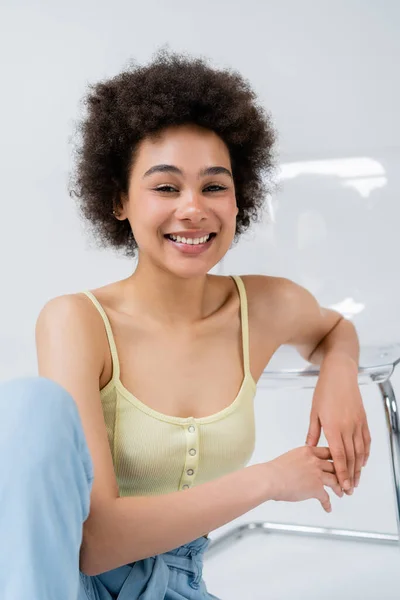 Cheerful african american model looking at camera near modern chair on grey background — Stock Photo
