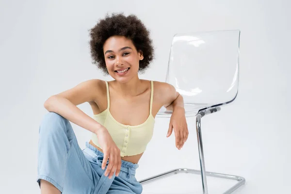 Curly african american woman sitting near modern chair on grey background — Stock Photo