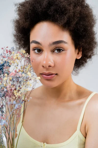 Portrait de femme afro-américaine avec maquillage naturel regardant la caméra près de fleurs de souffle de bébé isolé sur gris — Photo de stock