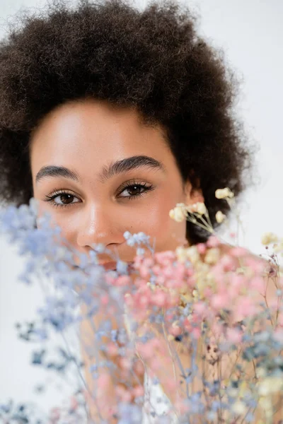 Femme afro-américaine regardant la caméra près de fleurs floues isolées sur gris — Photo de stock
