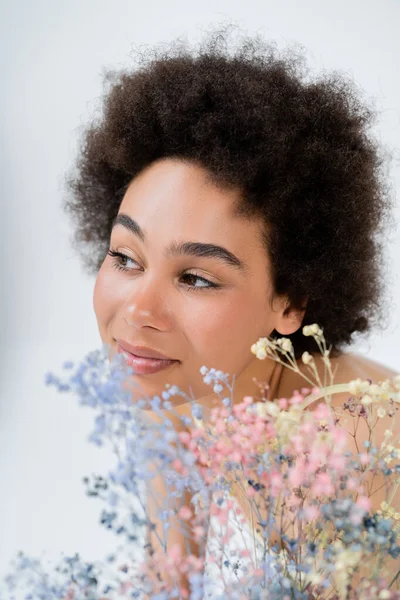 Positive african american woman looking away near baby breath flowers isolated on grey — Stock Photo