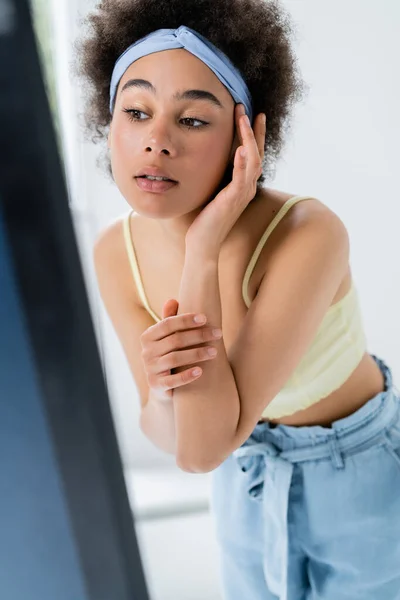 Junge afrikanisch-amerikanische Frau mit Stirnband blickt auf Spiegel auf grauem Hintergrund — Stockfoto