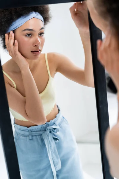 Bella donna afro-americana con fascia toccare faccia vicino specchio su sfondo grigio — Foto stock