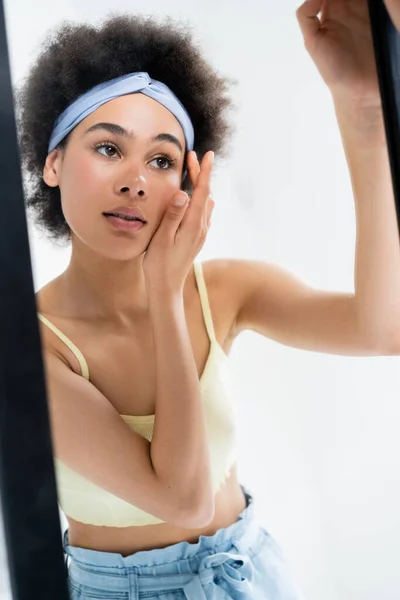 Junge Afroamerikanerin mit Stirnband berührt Gesicht in der Nähe Spiegel isoliert auf grau — Stockfoto