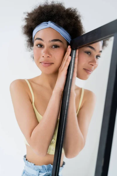 Lächelnde Afroamerikanerin mit Stirnband blickt in die Kamera in der Nähe von Spiegel isoliert auf grau — Stockfoto