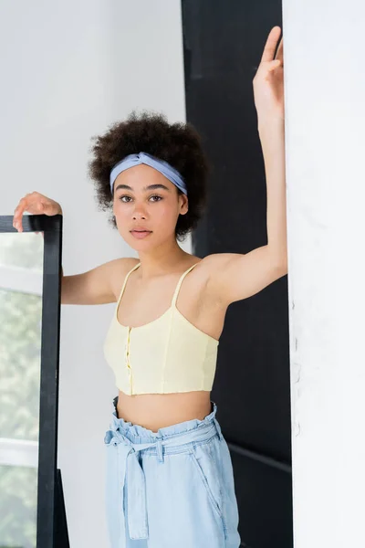 Curly african american woman in headband and top looking at camera near mirror on grey background — Stock Photo