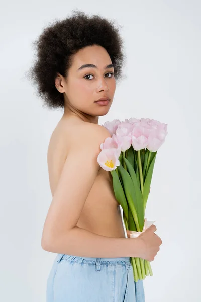 Jeune femme afro-américaine avec poitrine nue tenant des fleurs et regardant la caméra isolée sur gris — Photo de stock