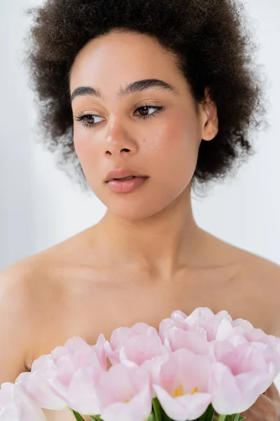 Portrait de jeune femme afro-américaine aux épaules nues tenant des tulipes roses isolées sur gris — Photo de stock