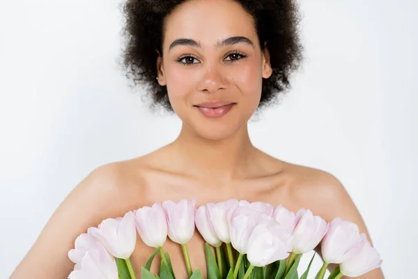 Mujer afroamericana sonriente con hombros desnudos sosteniendo tulipanes aislados en gris - foto de stock