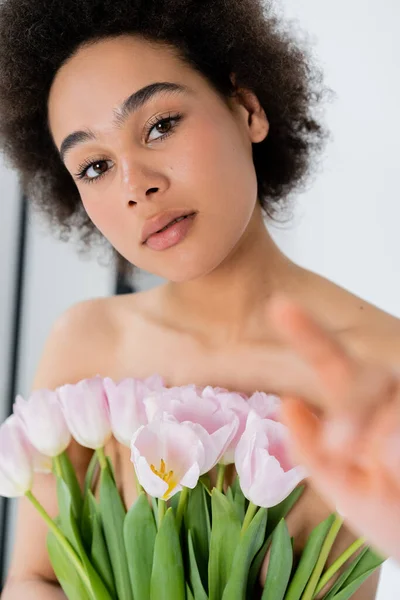 Mujer afroamericana con hombros desnudos sosteniendo tulipanes y señalando con el dedo sobre fondo gris - foto de stock