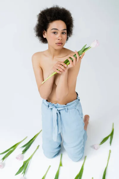 Кудрявая африканская американка с голыми плечами, держащая тюльпан на сером фоне — стоковое фото