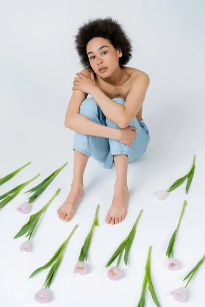 High angle view of african american woman with naked shoulders sitting near flowers on grey background — Stock Photo