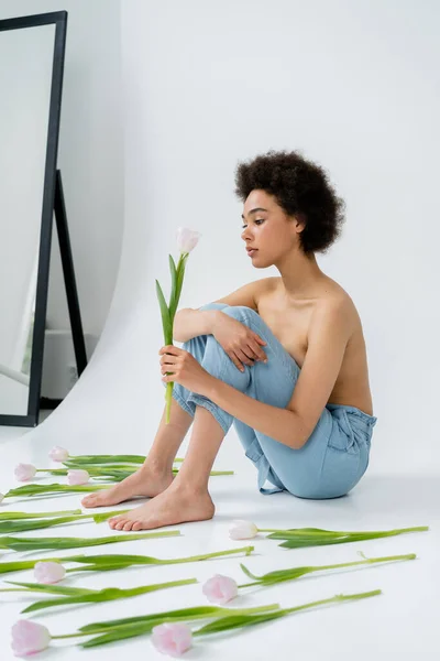 Afroamerikanerin mit nackter Brust hält Tulpenblume auf grauem Hintergrund — Stockfoto