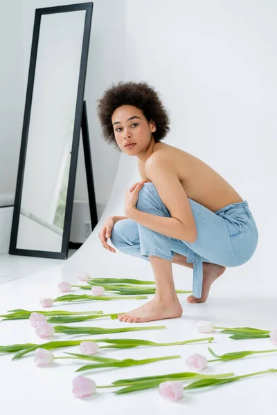 Young african american woman with naked shoulders looking at camera near tulips on grey background — Stock Photo