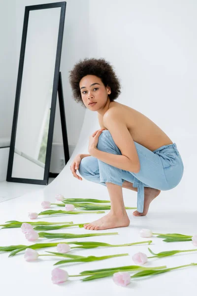 Jovem afro-americana com ombros nus olhando para a câmera perto de tulipas em fundo cinza — Fotografia de Stock