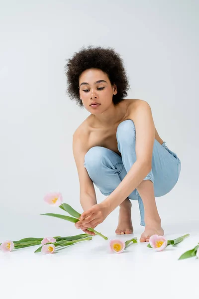 Barefoot african american woman with naked shoulders taking tulip on grey background — Stock Photo