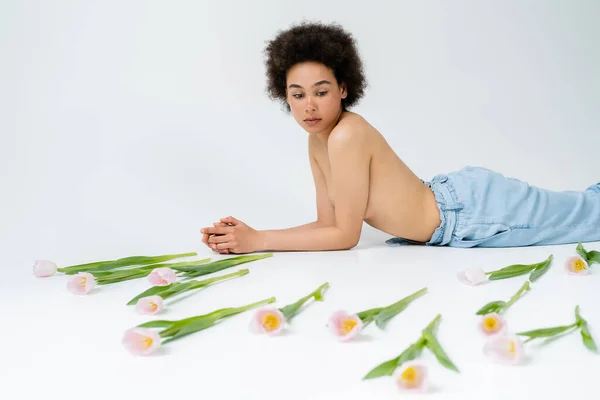 Afroamerikanerin mit nackter Brust liegt neben verschwommenen Tulpen auf grauem Hintergrund — Stockfoto