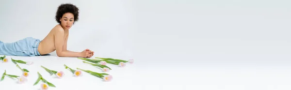 Donna afroamericana senza maglietta sdraiata vicino a fiori di tulipano su sfondo grigio, banner — Foto stock