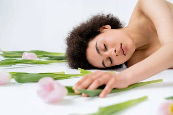 Young african american woman with naked shoulders lying near tulips on grey background — Stock Photo