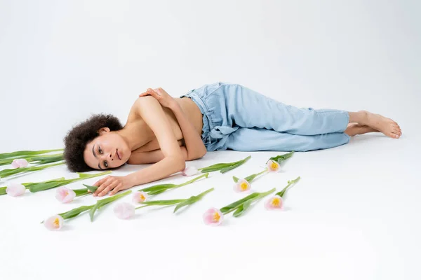 Американка без рубашки и босиком трогает цветок тюльпана, лежа на сером фоне. — стоковое фото
