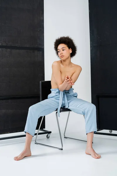 Shirtless african american woman posing on chair on grey and black background — Stock Photo