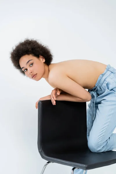 Femme afro-américaine torse nu en pantalon posant près d'une chaise isolée sur gris — Photo de stock