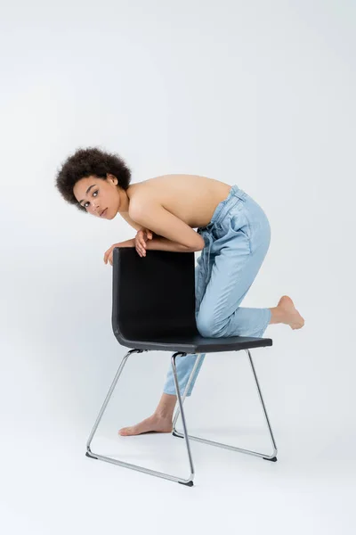 Афроамериканська жінка з босоніж і без сорочки стоїть біля сучасного крісла на сірому фоні. — стокове фото