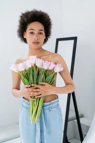 Mujer afroamericana sin camisa sosteniendo tulipanes cerca del pecho sobre fondo gris - foto de stock