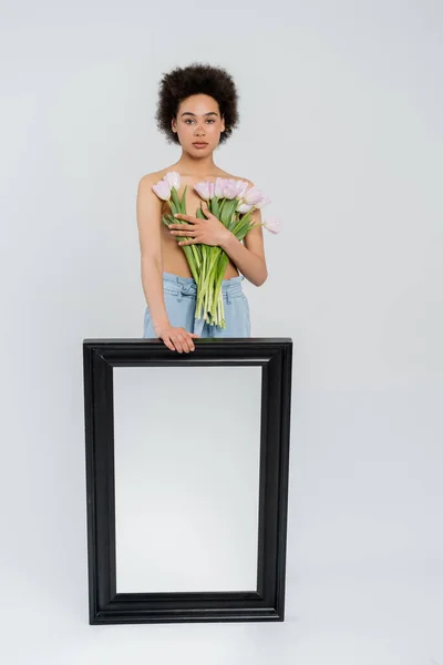 Африканская женщина без рубашки держит тюльпаны и зеркало на сером фоне — стоковое фото