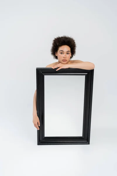 Африканская американка с голыми плечами, смотрящая в камеру возле зеркала на сером фоне — стоковое фото