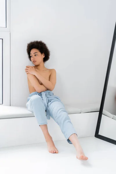 Mujer afroamericana sin camisa y descalza sentada sobre alféizar de ventana sobre fondo gris - foto de stock