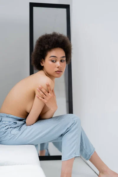 Без сорочки афроамериканська жінка торкається плеча, сидячи на підвіконні на сірому фоні — стокове фото