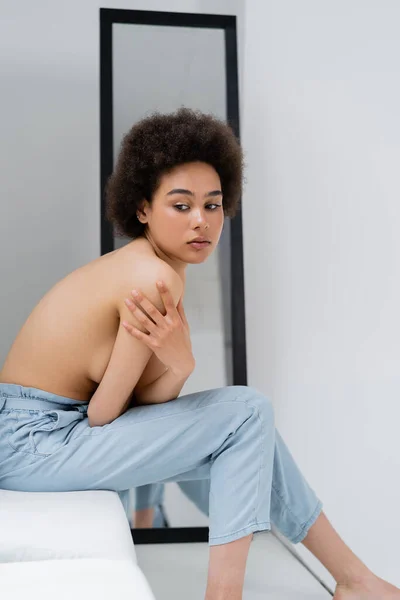 Shirtless african american woman touching shoulder while sitting on windowsill on grey background — Stock Photo