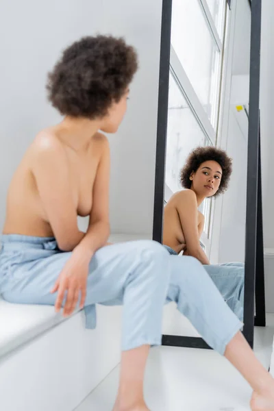 Mujer afroamericana sin camisa mirando el espejo mientras está sentada en el alféizar de la ventana sobre un fondo gris - foto de stock