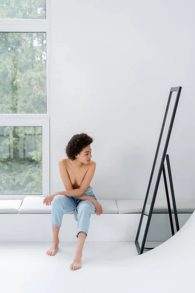 Hemdlose afrikanisch-amerikanische Frau sitzt neben Spiegel und Fenster auf grauem Hintergrund — Stockfoto