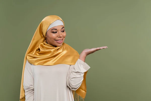 Positive multirassische Frau im Hijab zeigt mit der Hand isoliert auf grün — Stockfoto