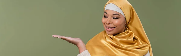Positiva donna multirazziale in hijab dorato che punta con mano isolata sul verde, striscione — Foto stock