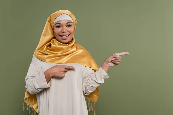 Despreocupada mujer multirracial en hiyab señalando con los dedos aislados en verde - foto de stock