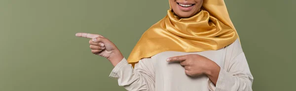 Ausgeschnittene Ansicht einer lächelnden multirassischen Frau im Hijab, die mit den Fingern auf grünem Banner zeigt — Stockfoto