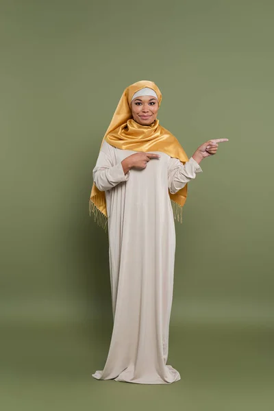 Full length of positive multiracial woman in hijab pointing with fingers on green background — Stock Photo