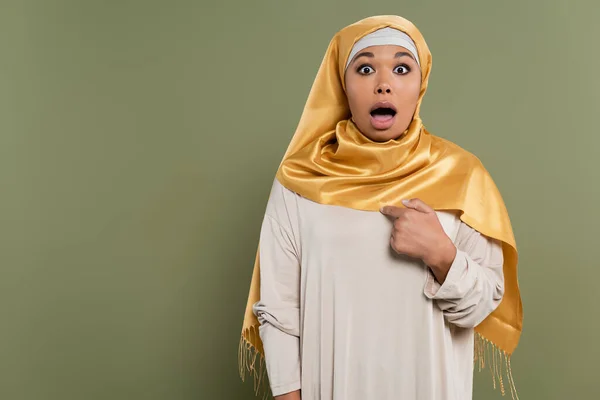 Impresionada mujer multirracial en hijab señalándose con el dedo sobre un fondo verde - foto de stock