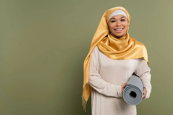 Smiling multiracial woman in hijab holding fitness mat on green background — Stock Photo