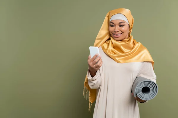 Smiling multiracial woman in hijab holding fitness mat and using smartphone on green background — Stock Photo